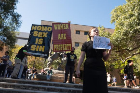 Two men are holding signs stating "Homo Sex is a Sin" and "Women are Property"